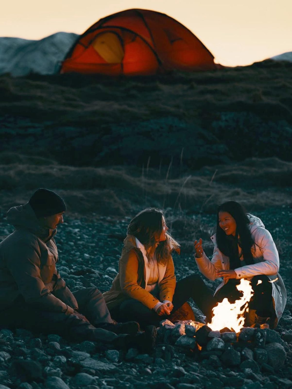 Friends around a campfire below a lit tent